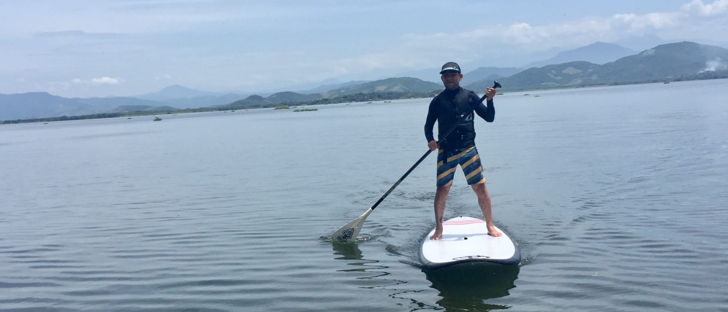 Hombre remando de pie sobre una tabla de paddle board en la laguna, con las montañas al fondo
