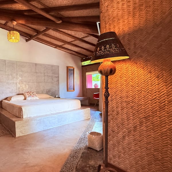 Habitación Petate con cama sobre base de concreto, muros de petate y lámpara de pie