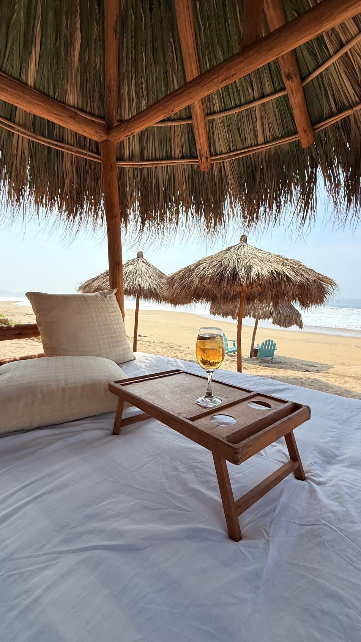 Camastro con cojines y una bebida bajo una palapa frente a la playa