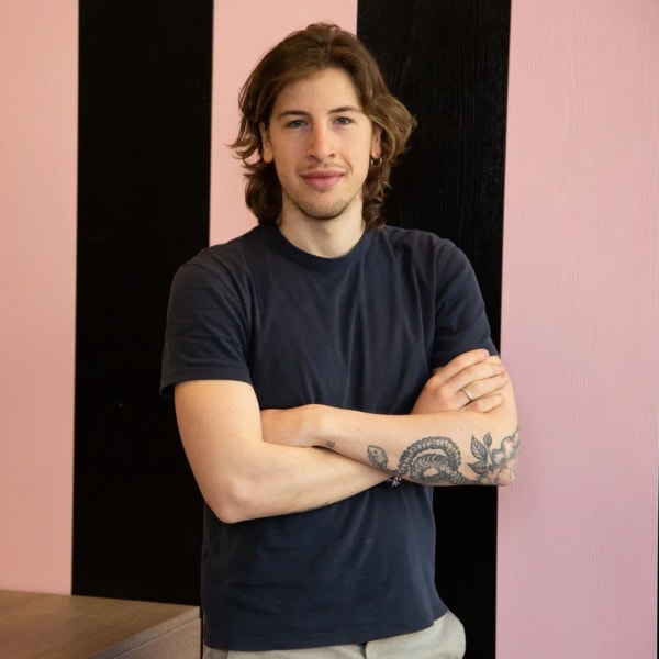 A young man with long brown hair and tattoos on his arms stands against a pink and black striped wall.