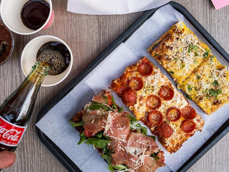 A tray of four rectangular Roman-style slices on a table, with a bottle of Coca-Cola being poured.