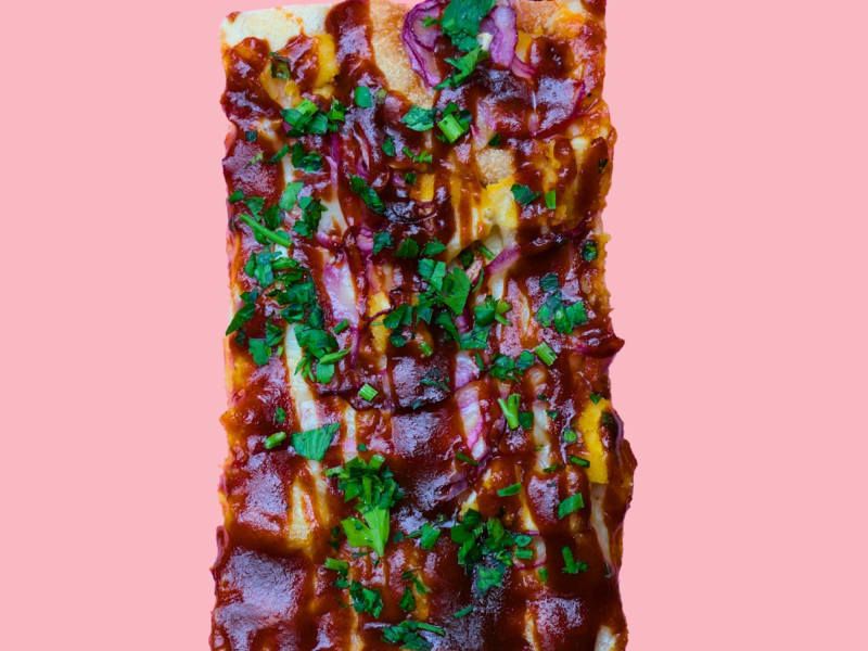 A rectangular slice of BBQ chicken pizza with red onions and cilantro on a pink background.