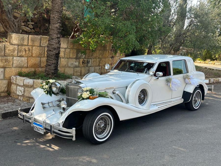 White 1978 Excalibur sedan with flowers on the grille, by a stone wall