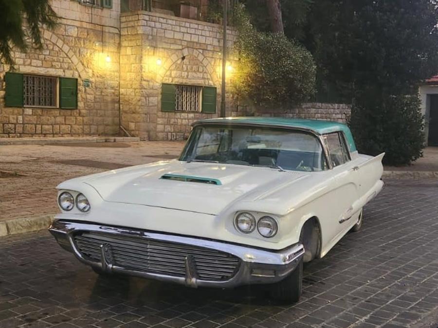 Cream 1960 Ford Thunderbird with a turquoise roof by a lit stone house at dusk
