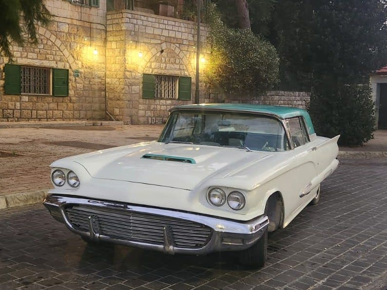 Cream 1960 Ford Thunderbird with a turquoise roof by a lit stone house at dusk