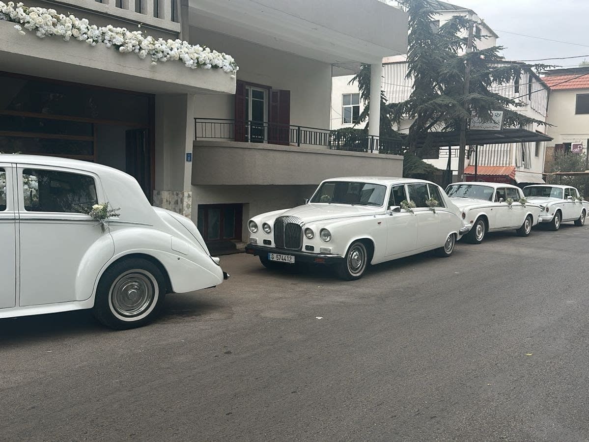 Row of white Daimler DS420 limousines outside a flower-decorated house