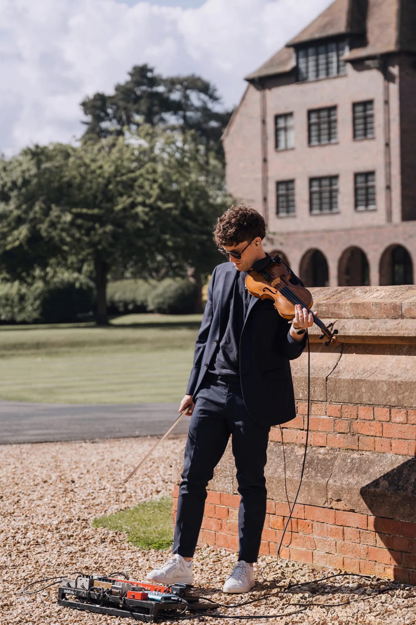 Joel Grainger playing violin outside a red-brick country house, his loop pedal on the gravel