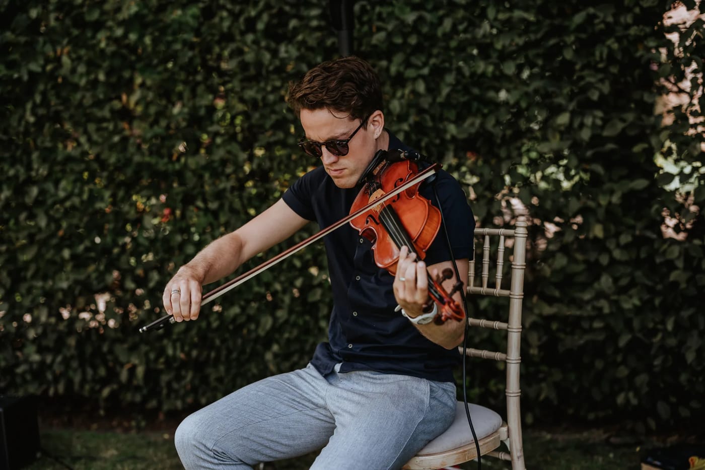 Joel Grainger seated with his violin beside an ivy hedge