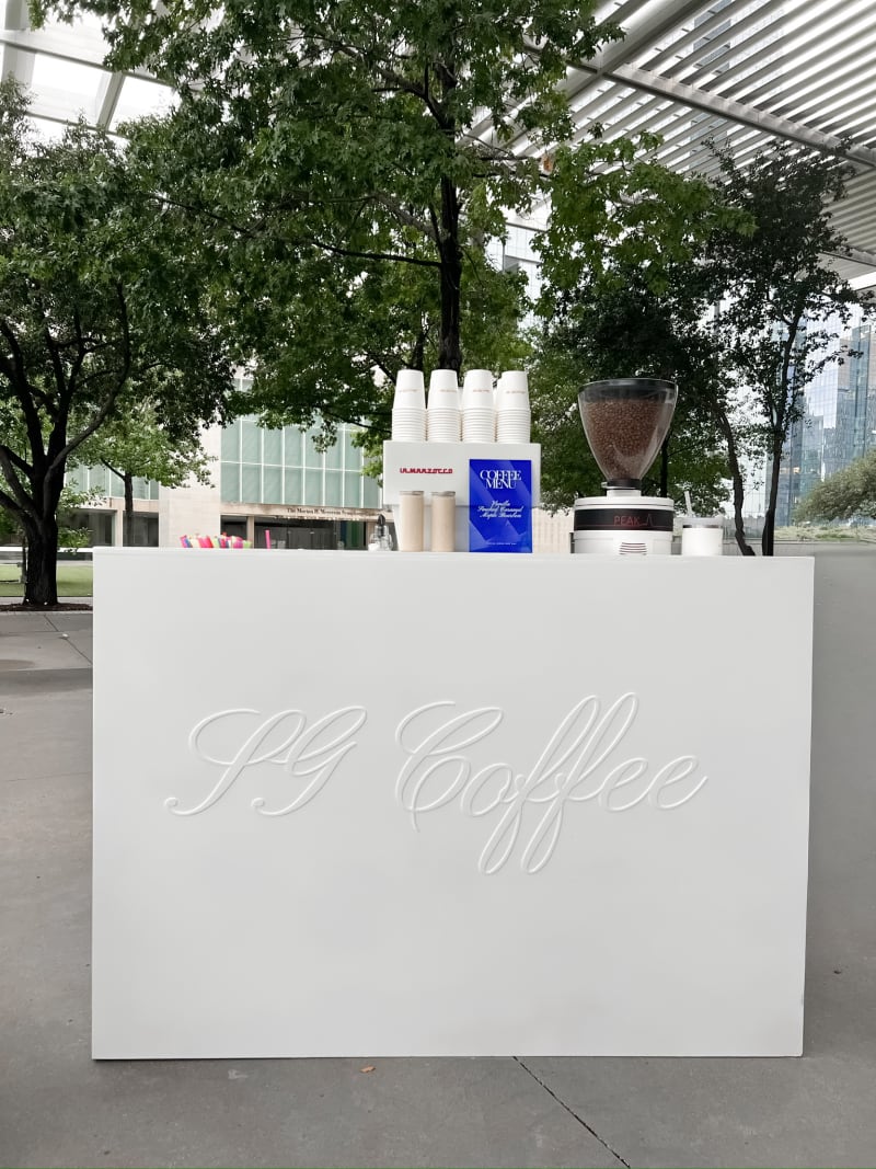 A white display stand with the text "PG Coffee" prominently displayed, set against a backdrop of lush greenery and a glass building in the distance.
