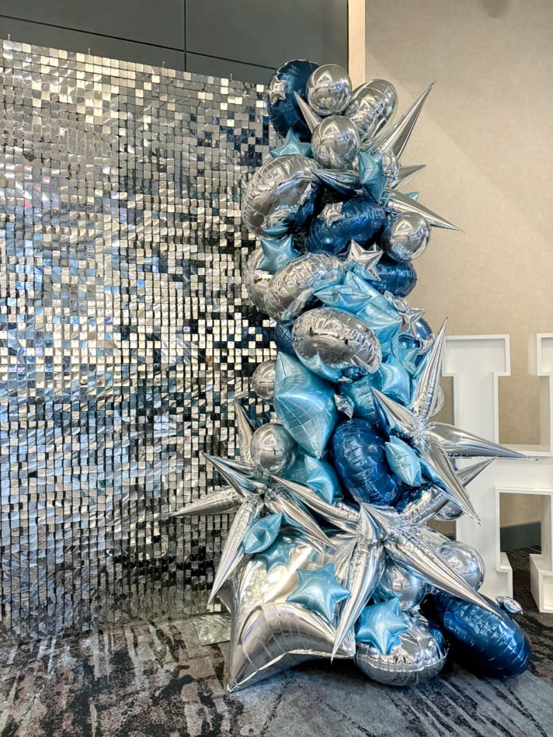 A striking, abstract sculpture composed of various shimmering, metallic elements in shades of blue and silver, set against a reflective, tiled wall backdrop.