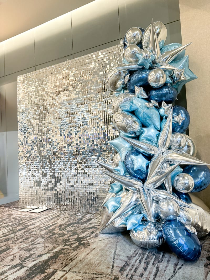 A large, abstract sculpture composed of shimmering blue and silver elements, set against a backdrop of a reflective, mosaic-like wall.