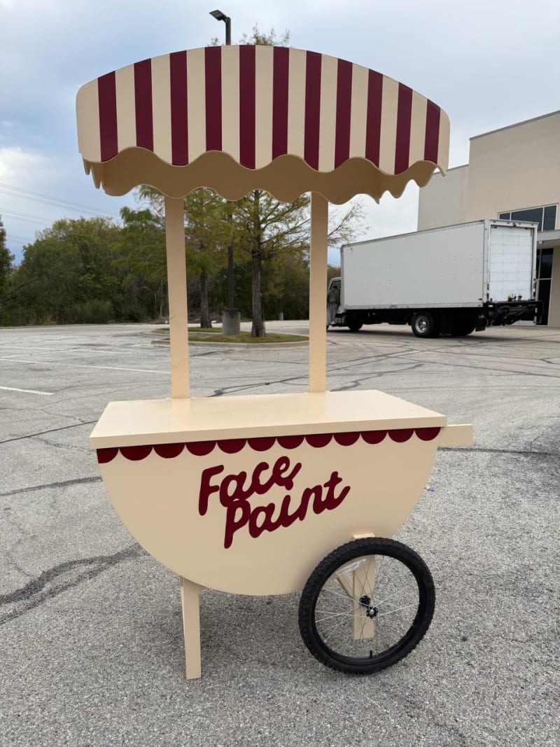 A colorful face paint cart stands in an empty parking lot, with a large truck visible in the background.