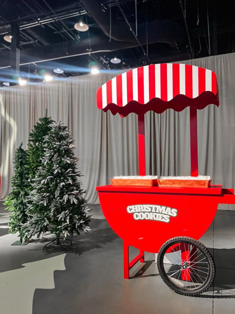A red and white striped Christmas cookie cart stands in front of a decorated Christmas tree in a dimly lit indoor setting.