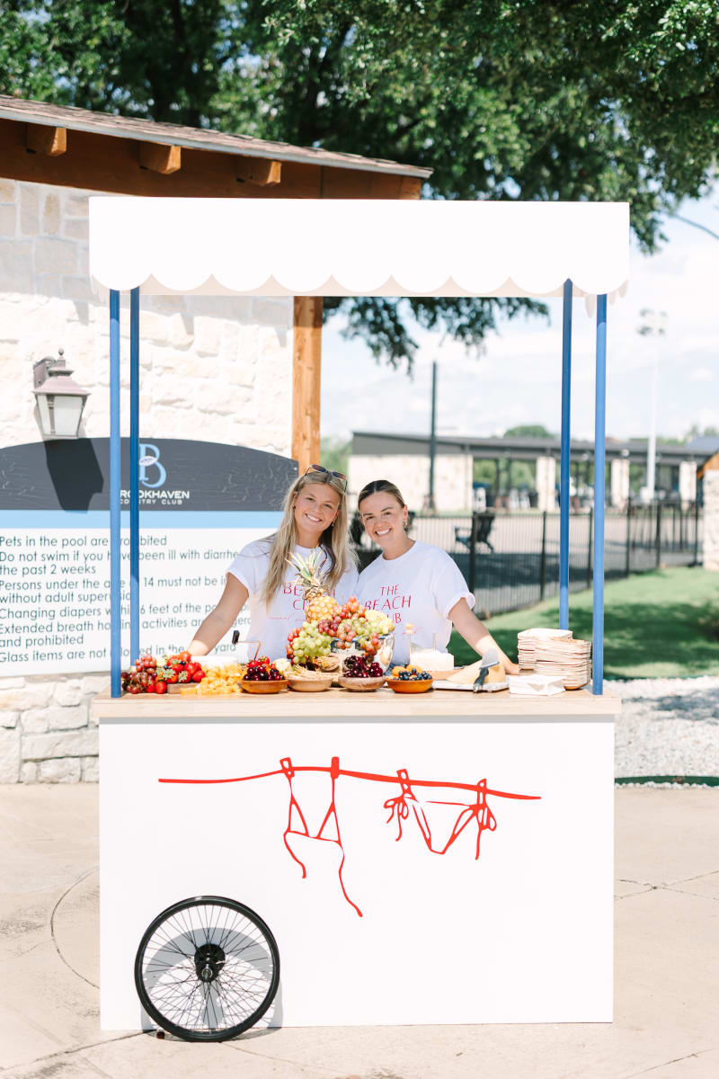 Two young women are standing behind a food cart, surrounded by a lush, green outdoor setting with trees in the background.
