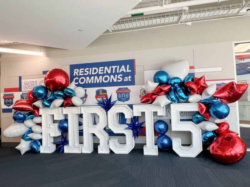 The image shows a large display of the text "FIRST 5" surrounded by various red and blue balloons, with a sign in the background that reads "RESIDENTIAL COMMONS".