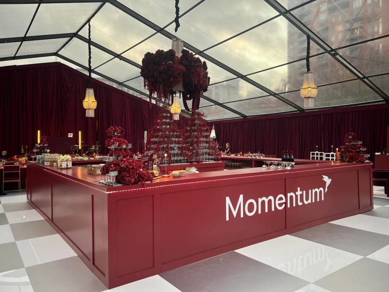 A modern, glass-enclosed event space with a large, red bar in the foreground displaying the word "Momentum", surrounded by various decorative elements and lighting fixtures.