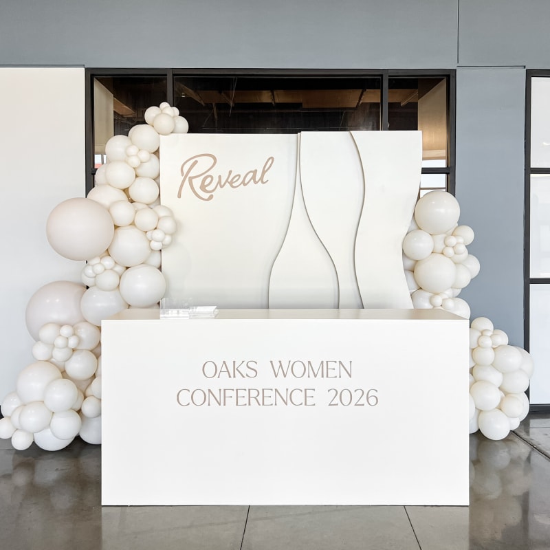 A large white display with the text "Reveal" and "OAKS WOMEN CONFERENCE 2026" stands in the foreground, surrounded by white balloon-like structures against a plain gray background.