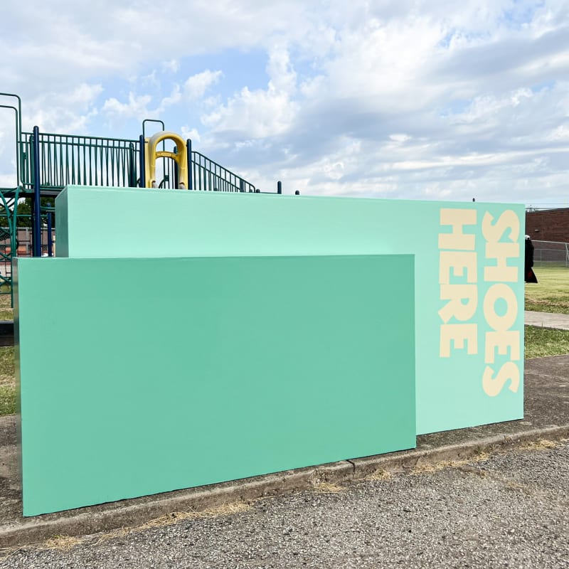 A large green wall with the text "HEROES" stands in the foreground, with a playground and fencing visible in the background against a cloudy sky.