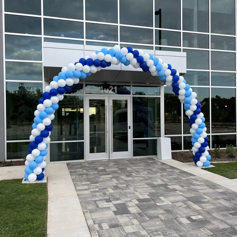 a blue and white balloon arch in front of a large glass building with a brick walkway in front of it