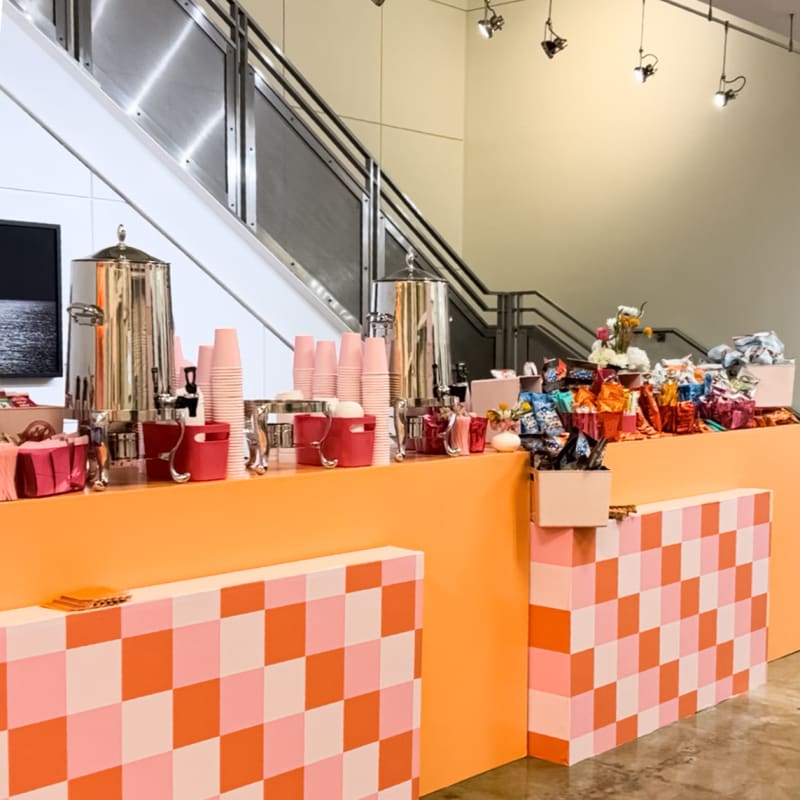 The image depicts a colorful and vibrant retail space with an orange and white checkered counter in the foreground, displaying various products and gifts. The background features glass walls, lighting fixtures, and a neutral-toned interior.