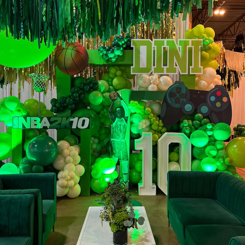 A vibrant and festive green-themed party setup, featuring various decorative elements such as hanging streamers, balloons, and a large "DINI" sign, along with gaming-related props like a controller, creating an immersive and engaging atmosphere.