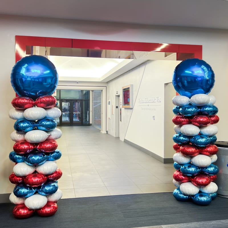 The image depicts a decorative archway made of red, white, and blue balloons, creating a festive and patriotic atmosphere. The background shows a hallway with white walls and a doorway visible.