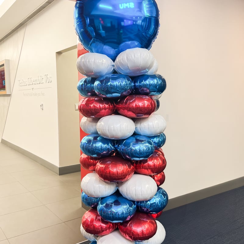 A tall, colorful tower of inflated balloons in various shades of blue, red, and white stands in a room with a tiled floor and white walls.
