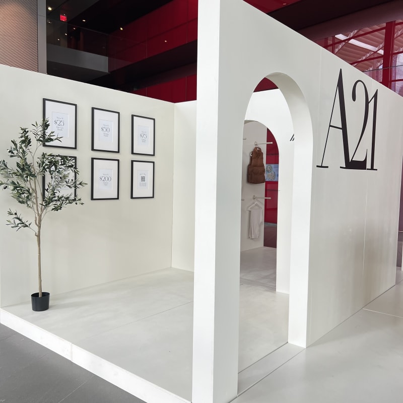 A modern, minimalist exhibition space with white walls, framed artwork, and a large "A21" sign on the wall.