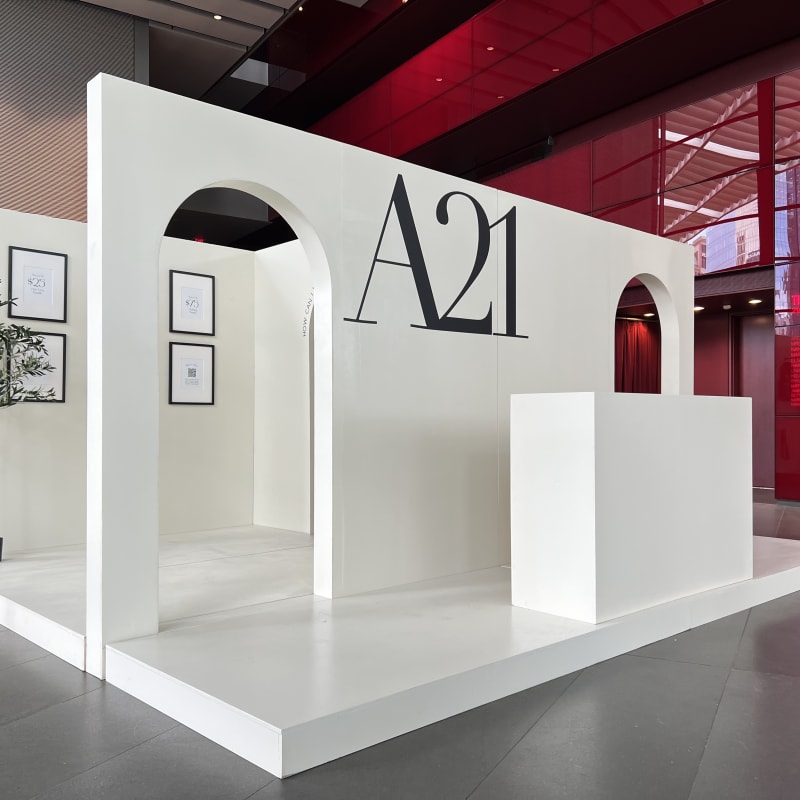 A minimalist, white exhibition booth with large black lettering "A21" and various architectural elements, set against a red-tinted background.