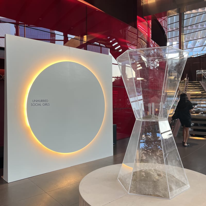 A large, illuminated circular display stands in the foreground, surrounded by a modern, minimalist interior with red lighting and architectural elements.