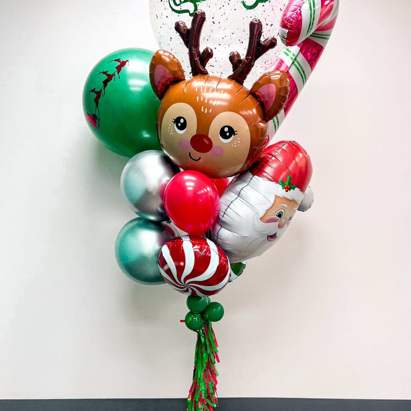 The image shows a colorful bouquet of Christmas-themed balloons, including a reindeer-shaped balloon, candy cane-shaped balloons, and a "Merry Christmas" balloon, all set against a plain white wall and wooden floor.