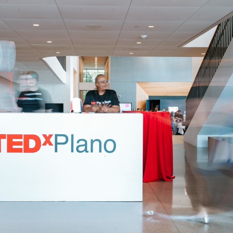 A reception desk with the TEDxPlano logo, surrounded by people in a modern, well-lit interior space.