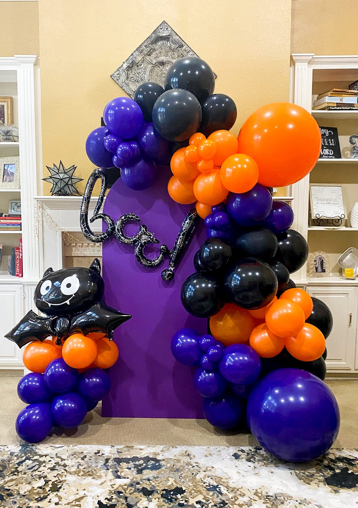 A vibrant display of purple, black, and orange balloons arranged in an abstract, sculptural form on a kitchen counter, with shelves and decor visible in the background.