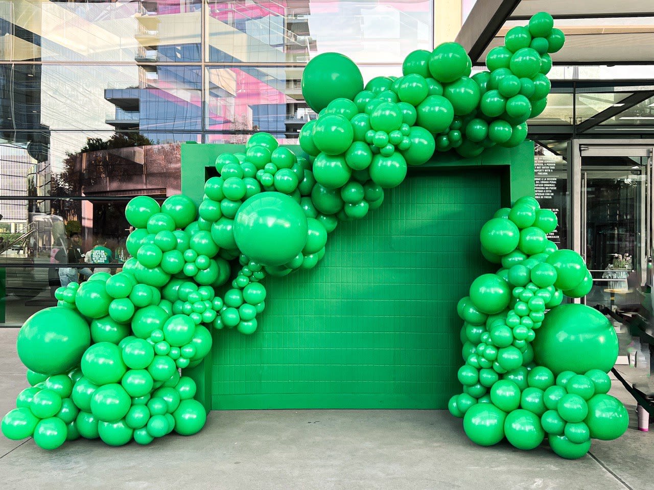 a large number of green balloons in front of a green wall with a green building in the back ground