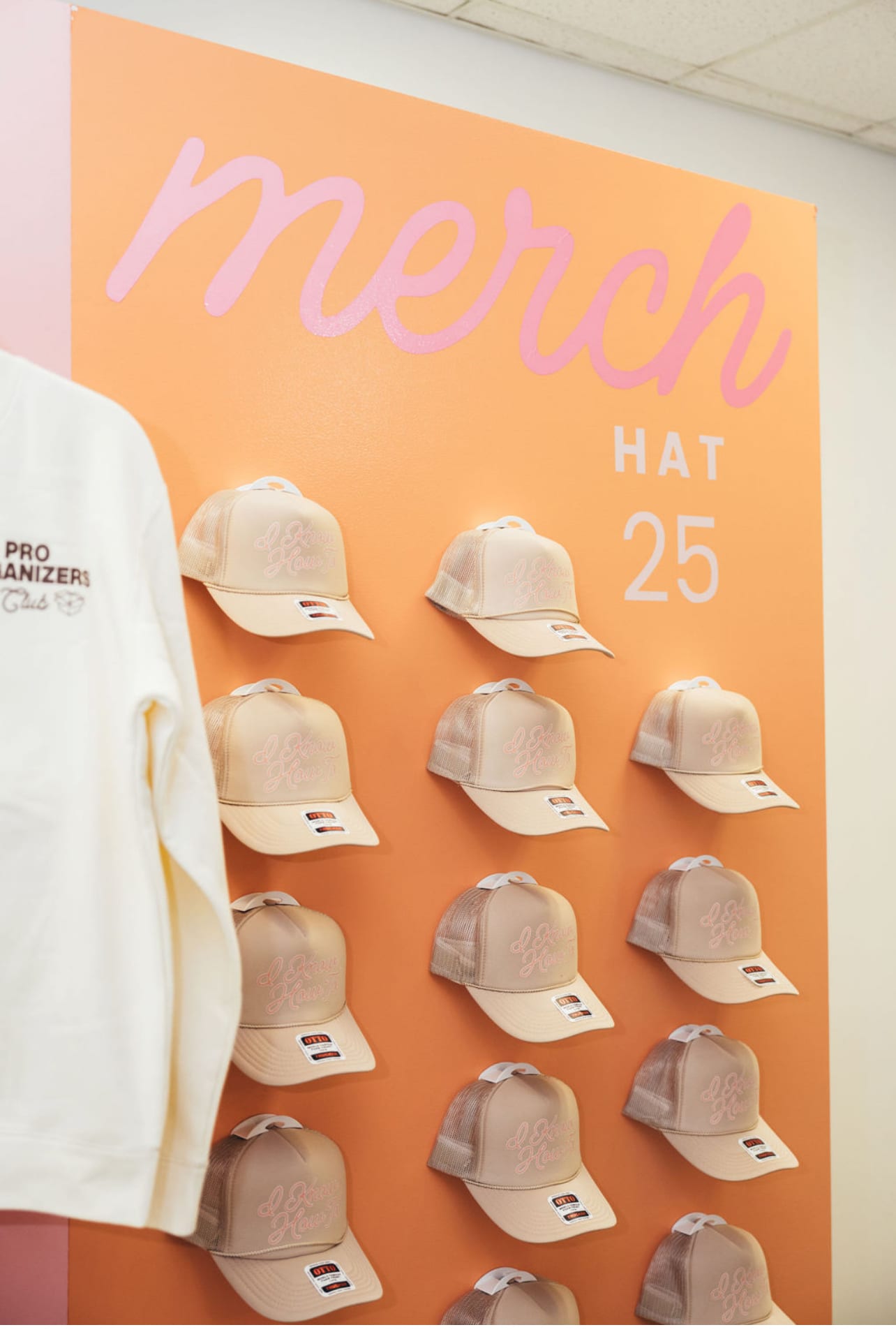 The image displays a display of white baseball caps or hats with the text "Merch Hat 25" prominently displayed on an orange background.