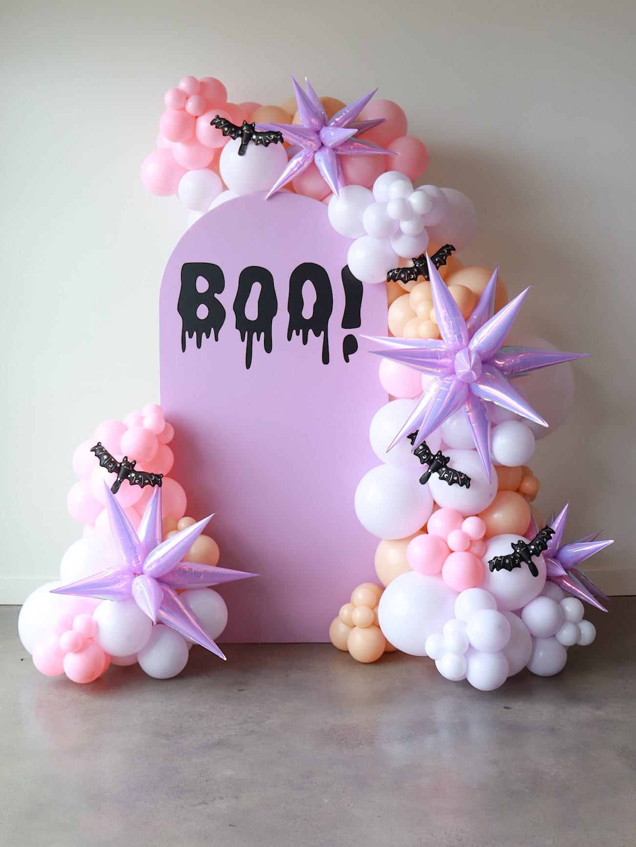 A pink and purple decorative display featuring the word "BOO!" surrounded by fluffy white and pink clouds, with purple star-shaped flowers and black bats.