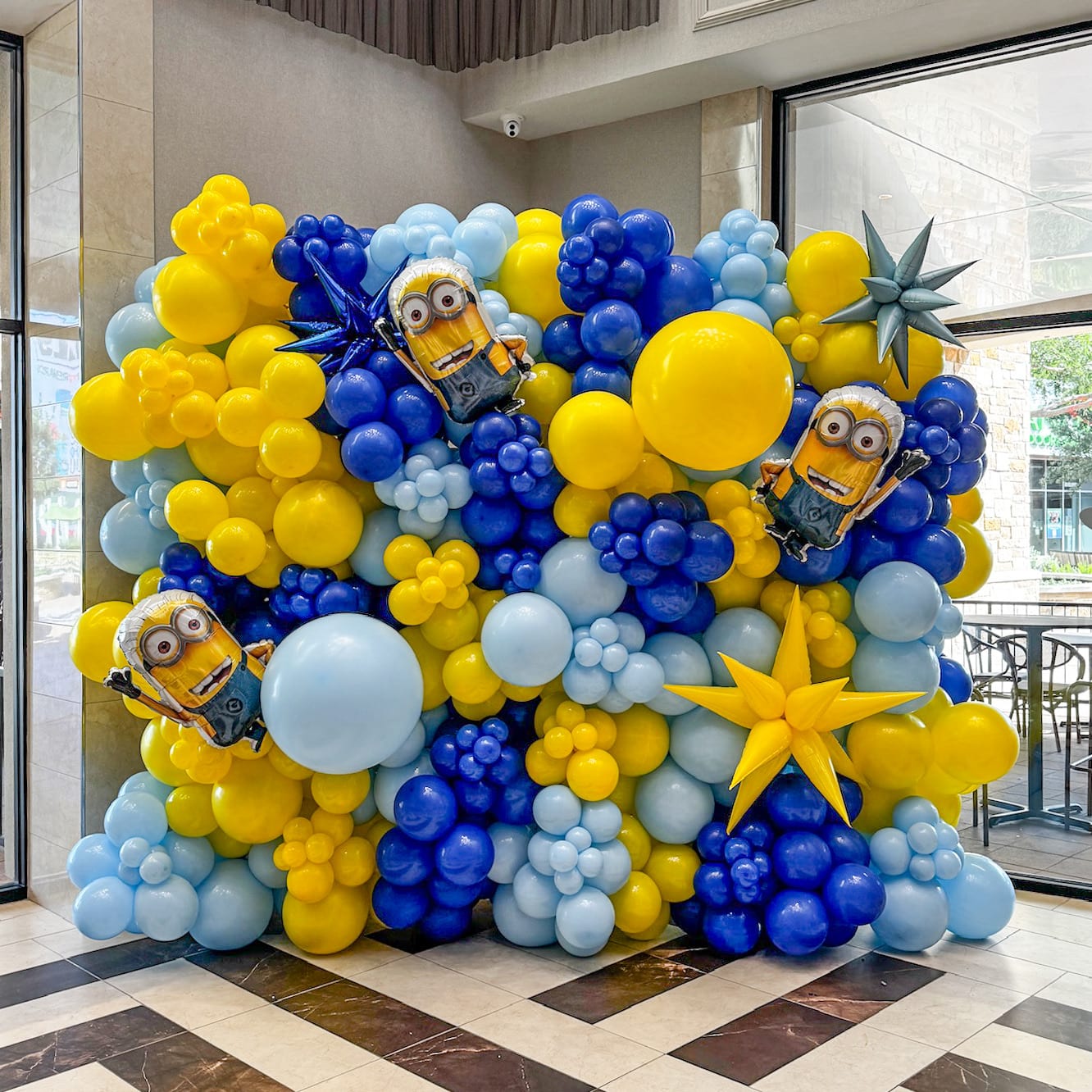 A vibrant and whimsical balloon display featuring a variety of yellow, blue, and white balloons, along with several Minion characters, adorns the interior space, creating a festive and playful atmosphere.