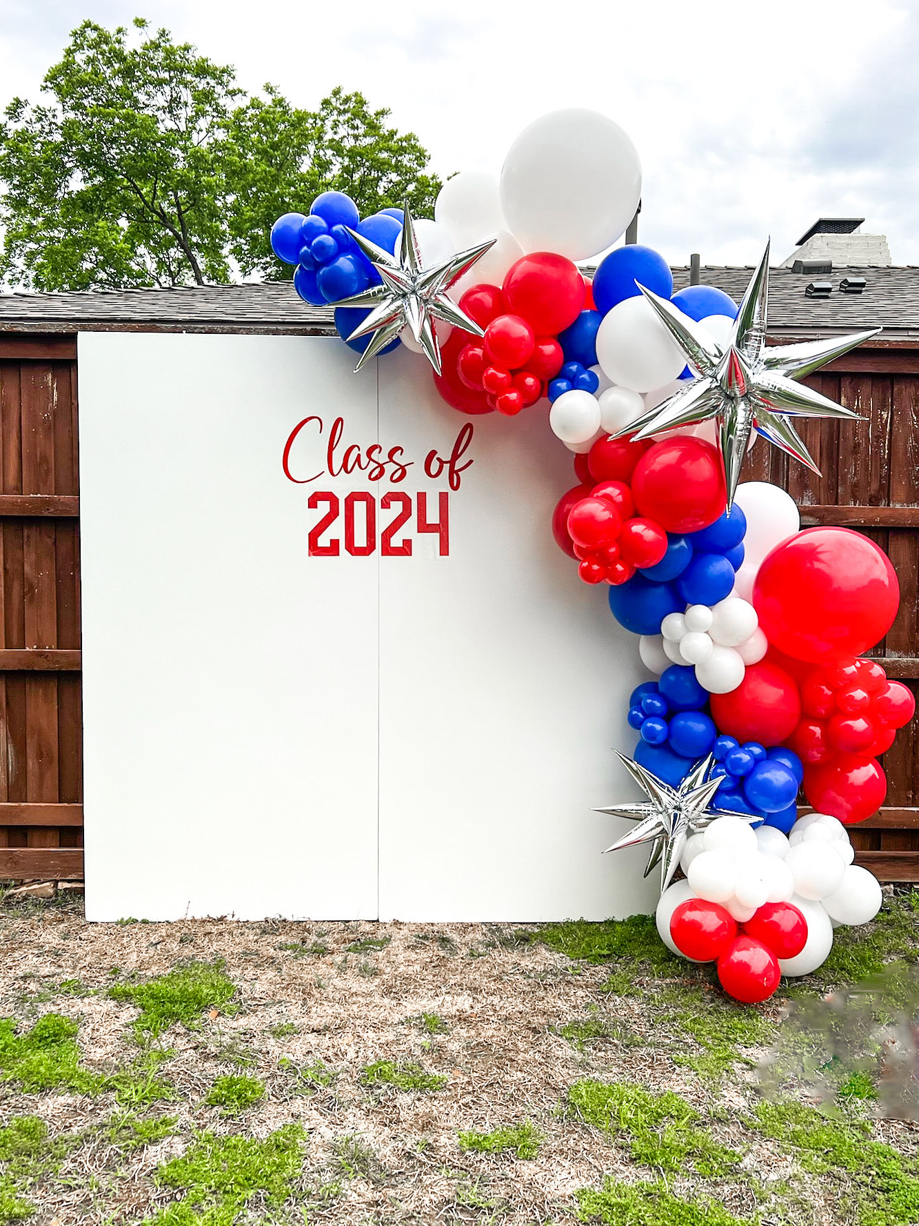 A festive backdrop with red, white, and blue balloons, stars, and the text "Class of 2024" against a wooden structure surrounded by lush greenery.