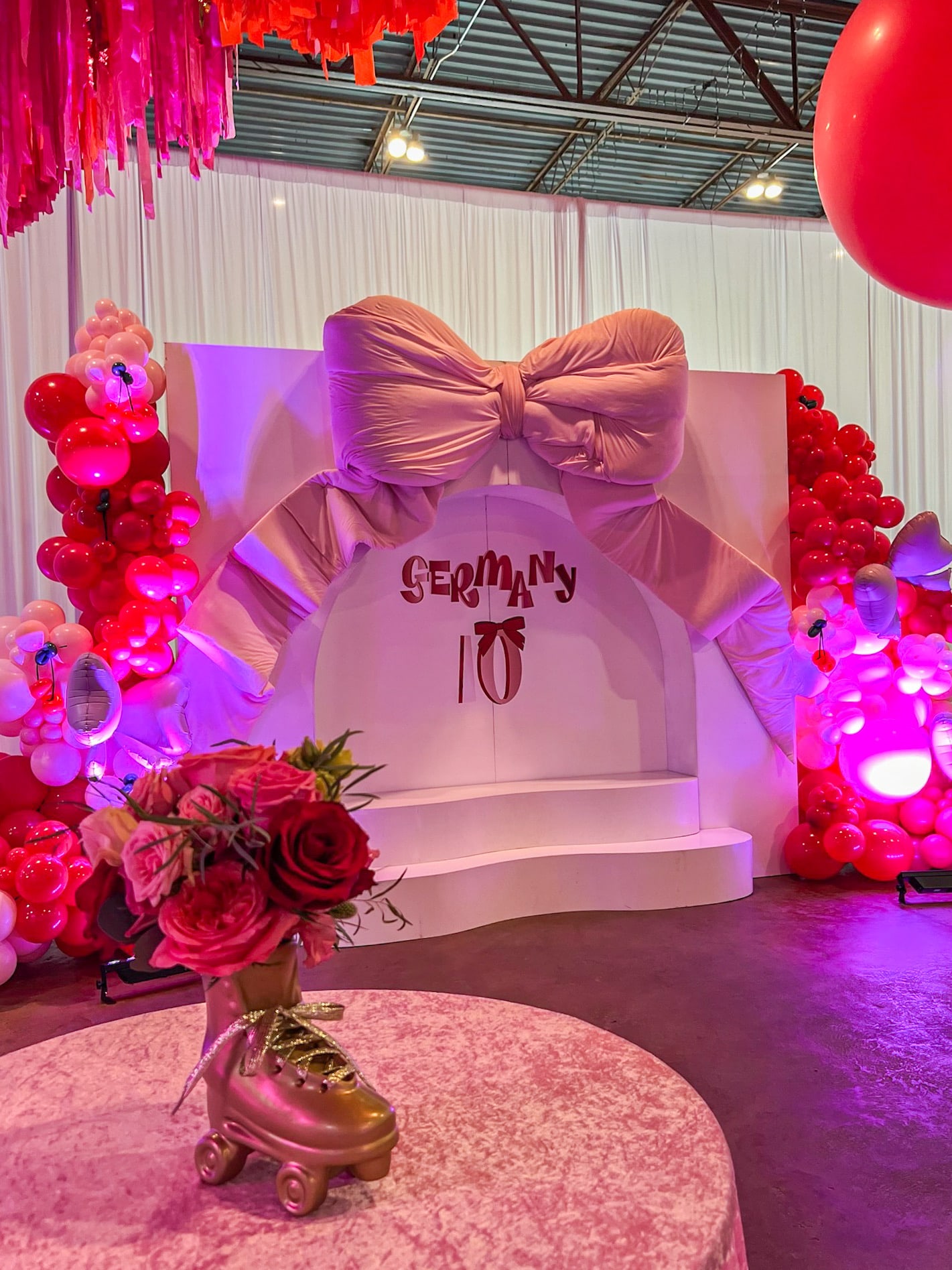 A lavishly decorated event space with a large white bow-adorned backdrop, surrounded by vibrant red and pink floral arrangements, glowing lights, and a pair of roller skates on a circular platform in the foreground.