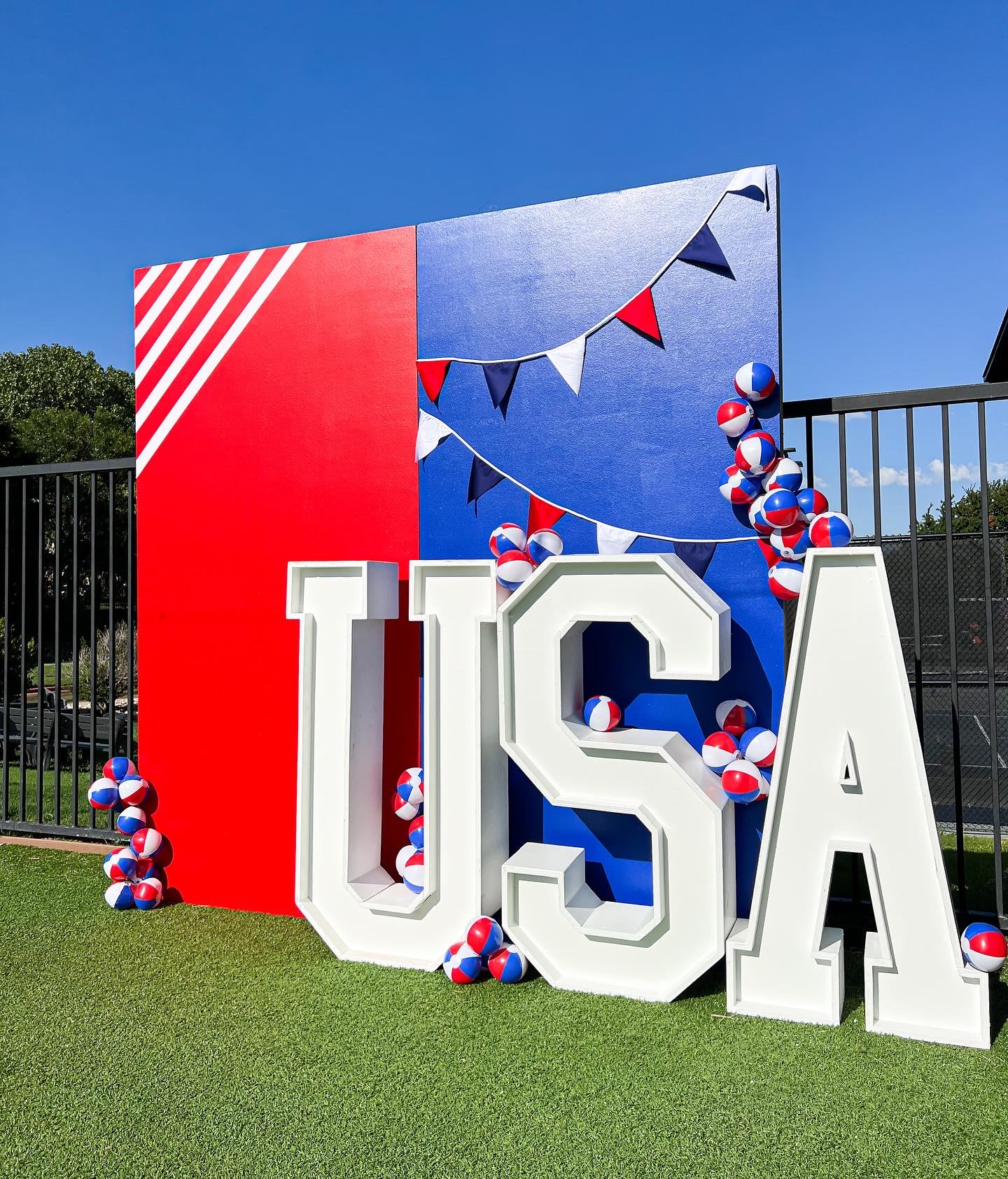 A large, colorful display featuring the letters "USA" surrounded by red, white, and blue decorations, set against a bright blue sky with trees in the background.