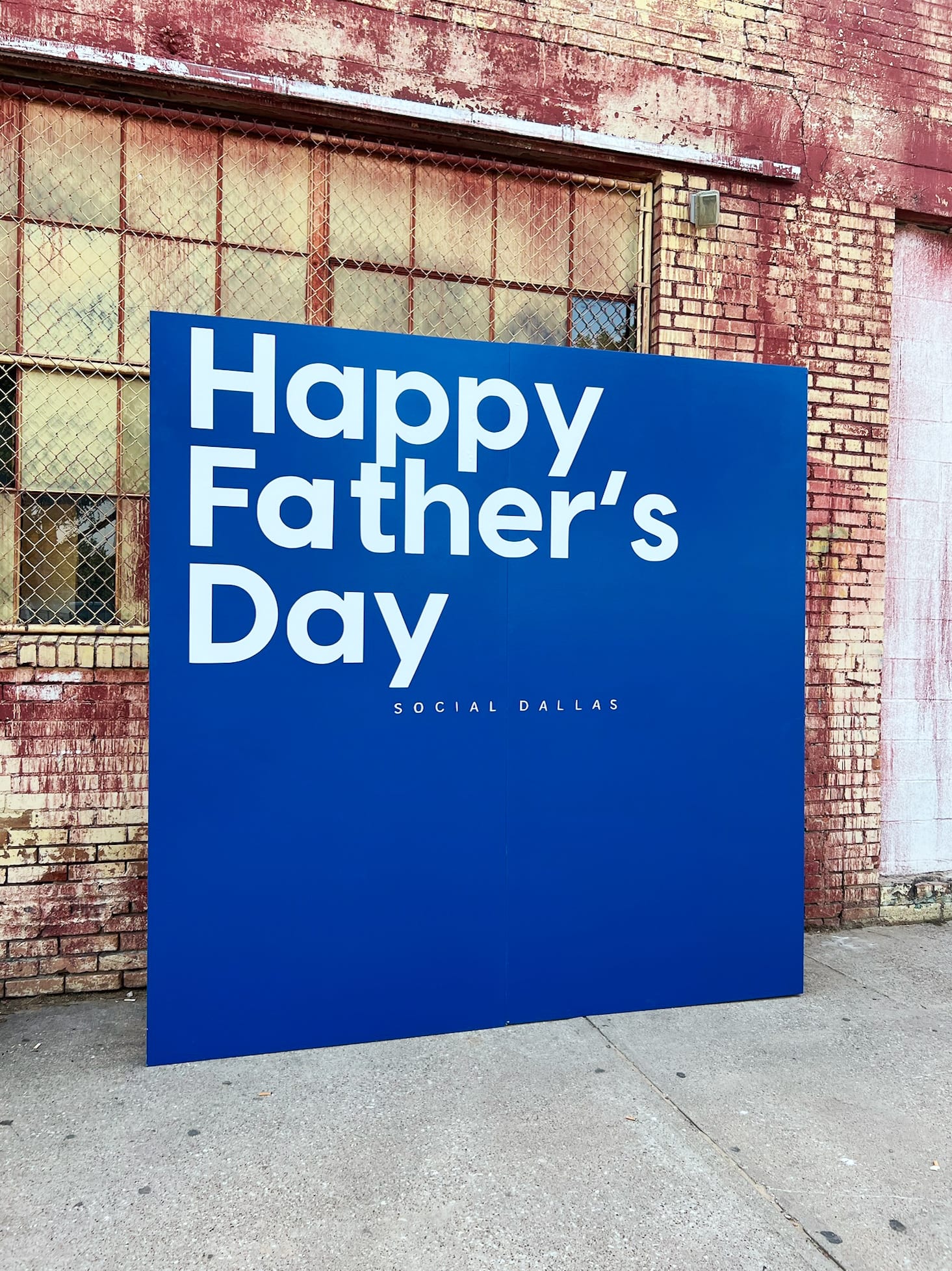 A large blue sign with the text "Happy Father's Day" is displayed in front of a brick building with a metal grate covering the windows.