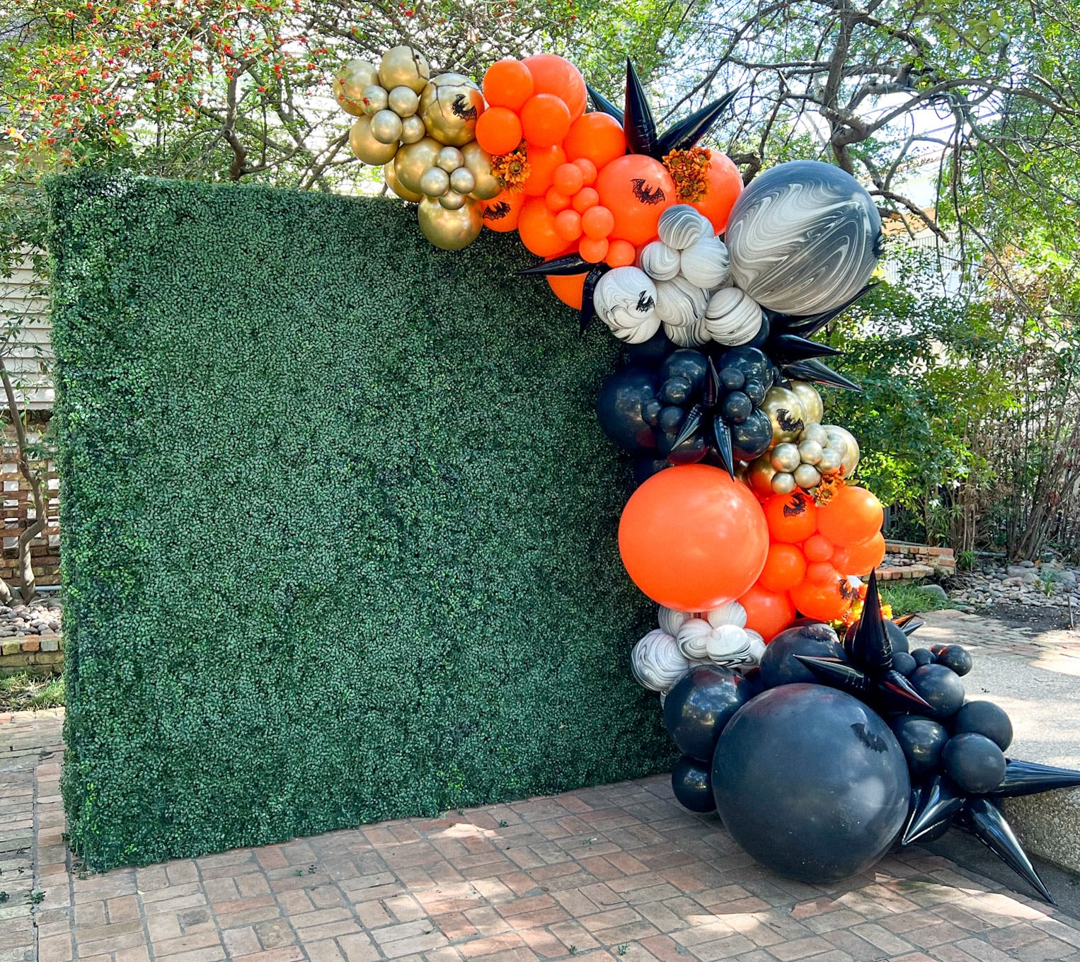 A vibrant and whimsical balloon display featuring a variety of colorful balloons, including orange, black, and white, arranged in an artistic manner against a backdrop of lush greenery and a brick wall.