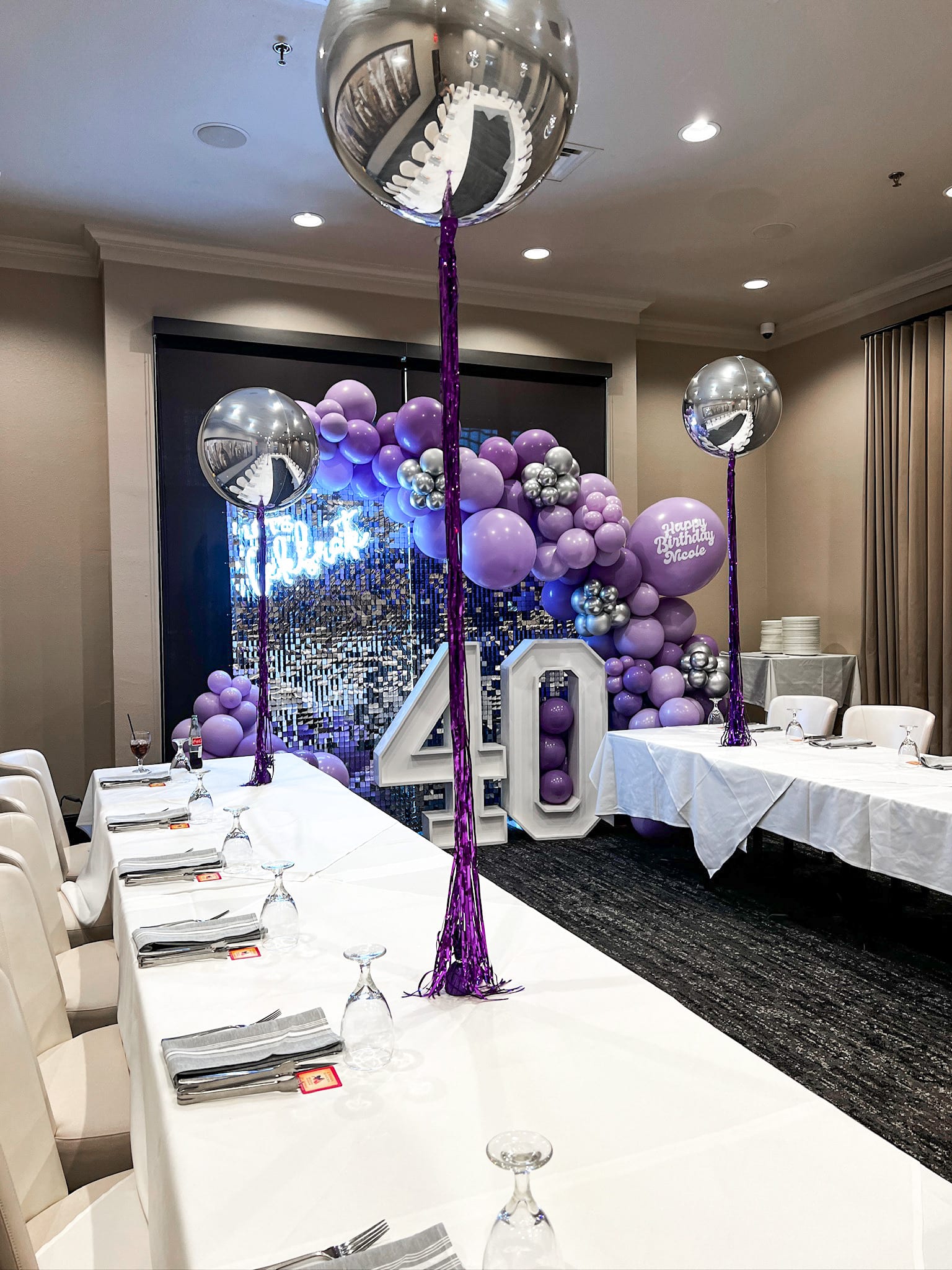 A festive event space with a large display of purple balloons, a mirrored disco ball, and a table set for a celebration, likely a 40th birthday party.