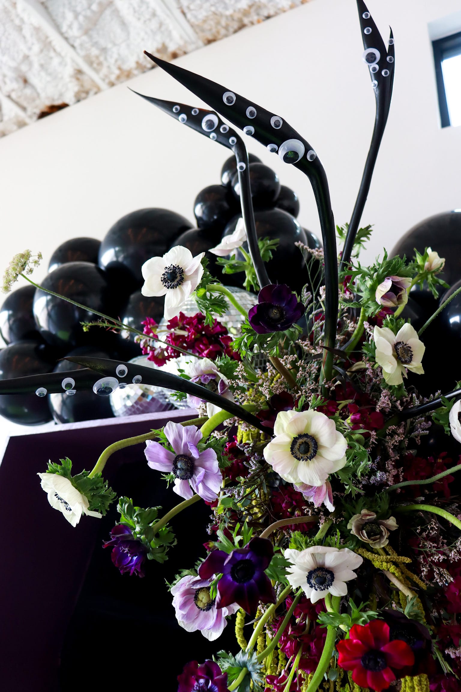 A vibrant bouquet of colorful flowers, including white anemones, purple pansies, and dark berries, sits against a backdrop of a rustic wooden surface.