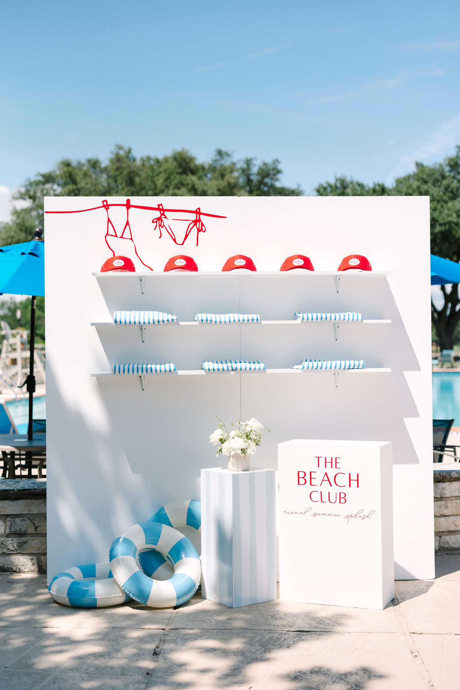 A white display stand with red lifesaving equipment and a sign reading "The Beach Club" stands in the foreground, with a scenic outdoor setting featuring trees and a blue sky visible in the background.