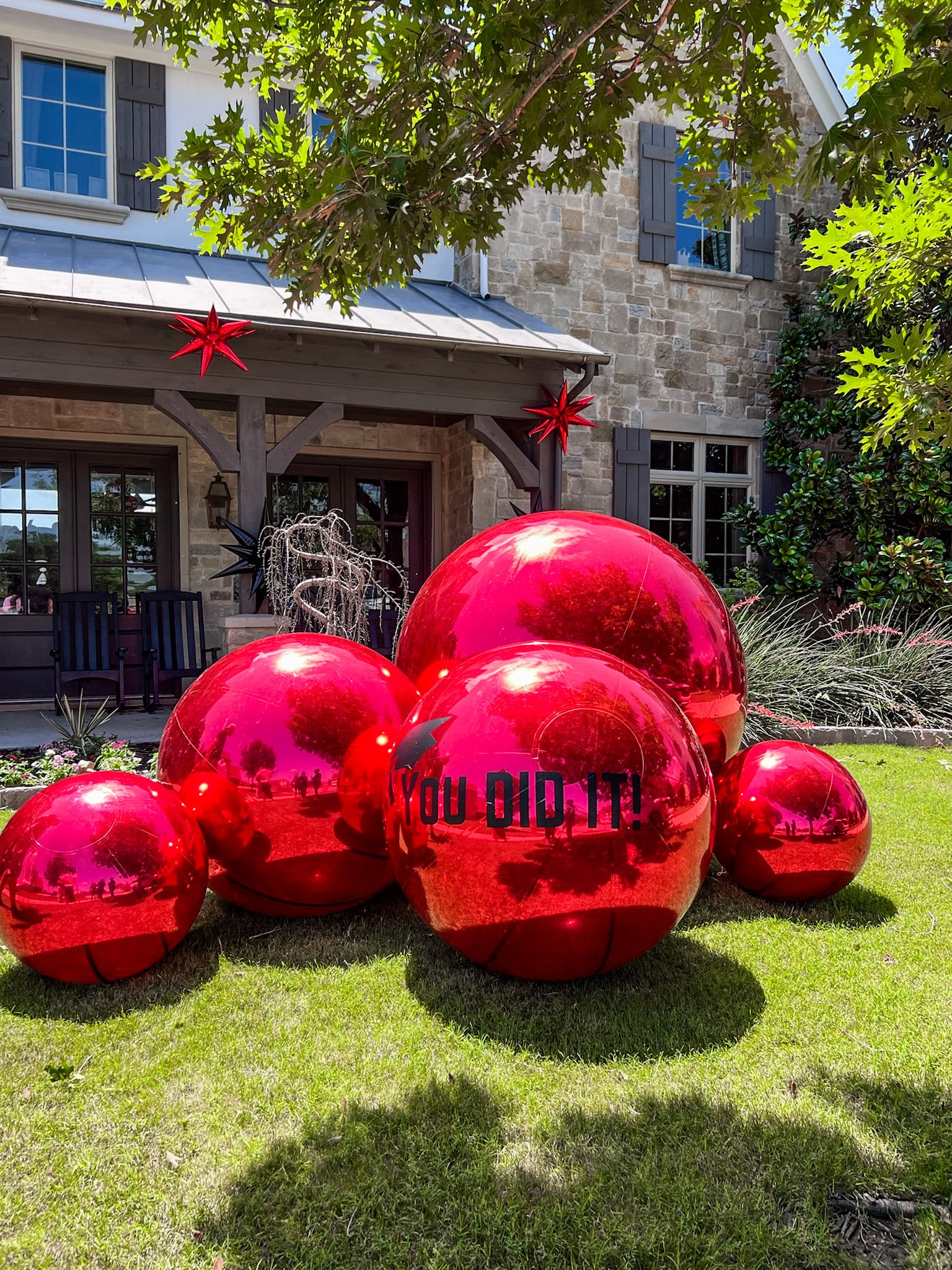 Large, shiny red spherical sculptures are displayed on a grassy lawn in front of a stone building with windows and a covered porch.