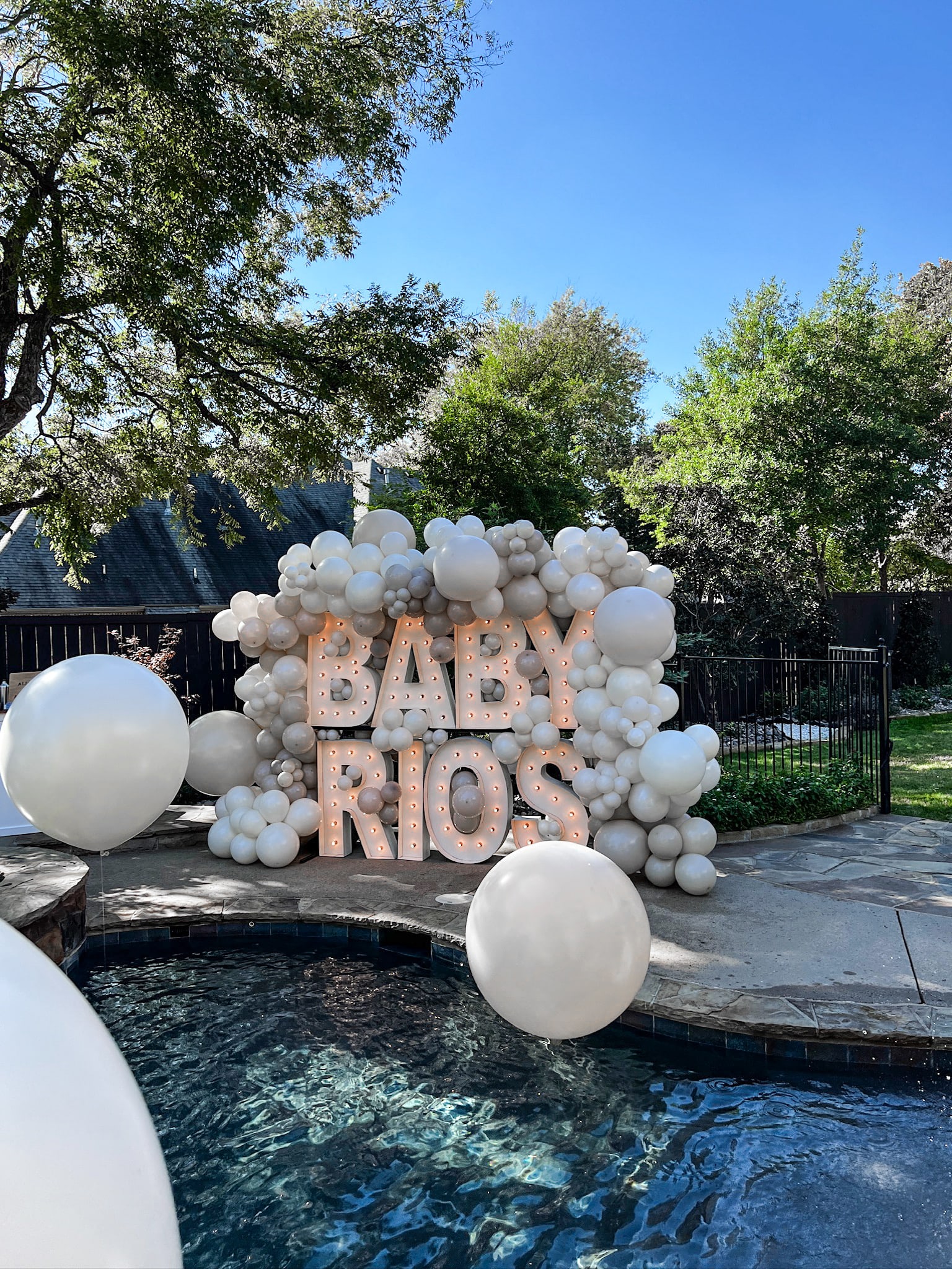A large display of white balloons forms the text "BABY RIOS" against a backdrop of lush greenery and a swimming pool.