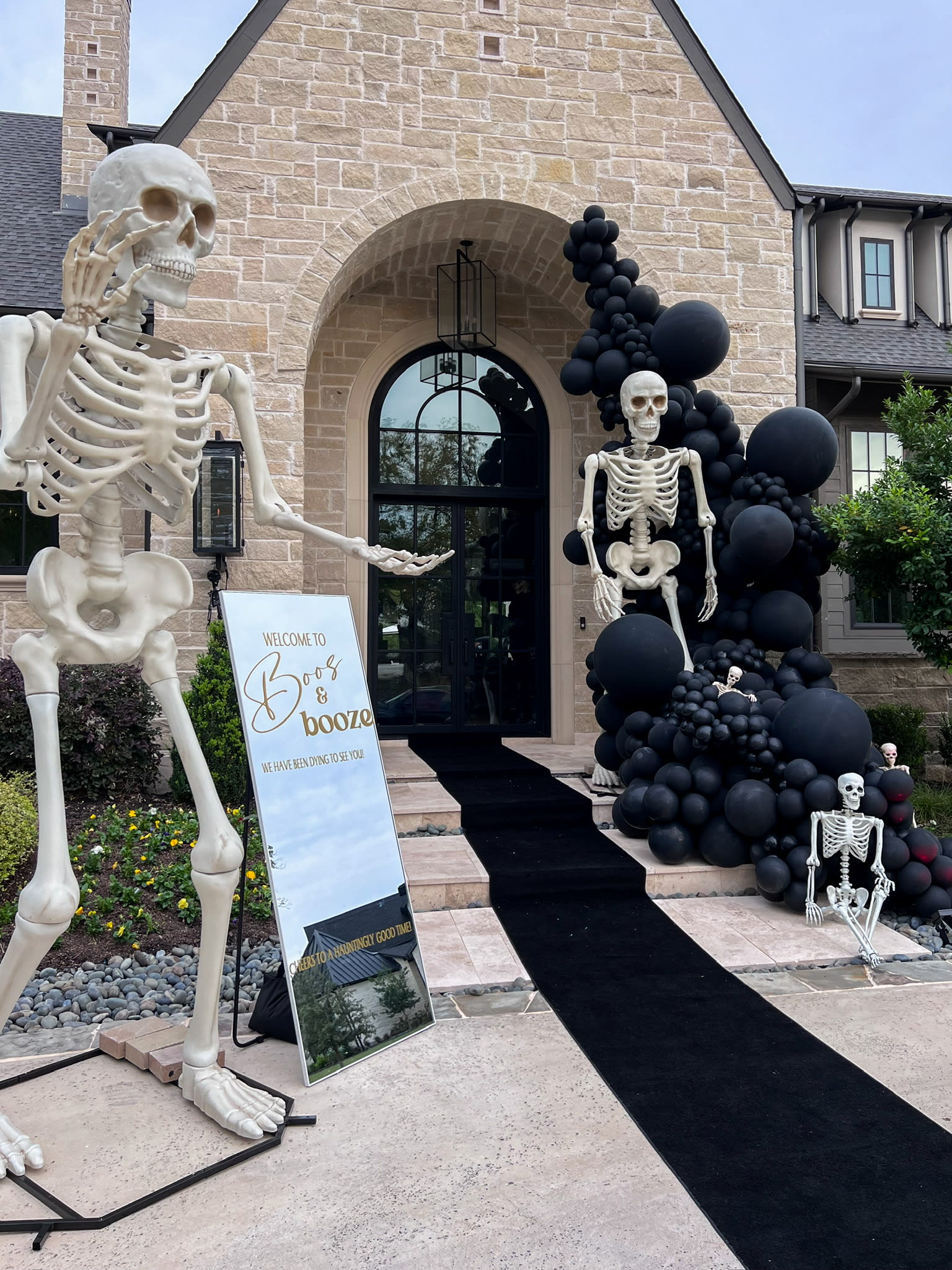 A large skeleton figure stands in the foreground, with a sign advertising "Spooky Books" nearby. In the background, a brick building with an arched entryway is surrounded by black balloon decorations, creating a spooky and festive Halloween atmosphere.