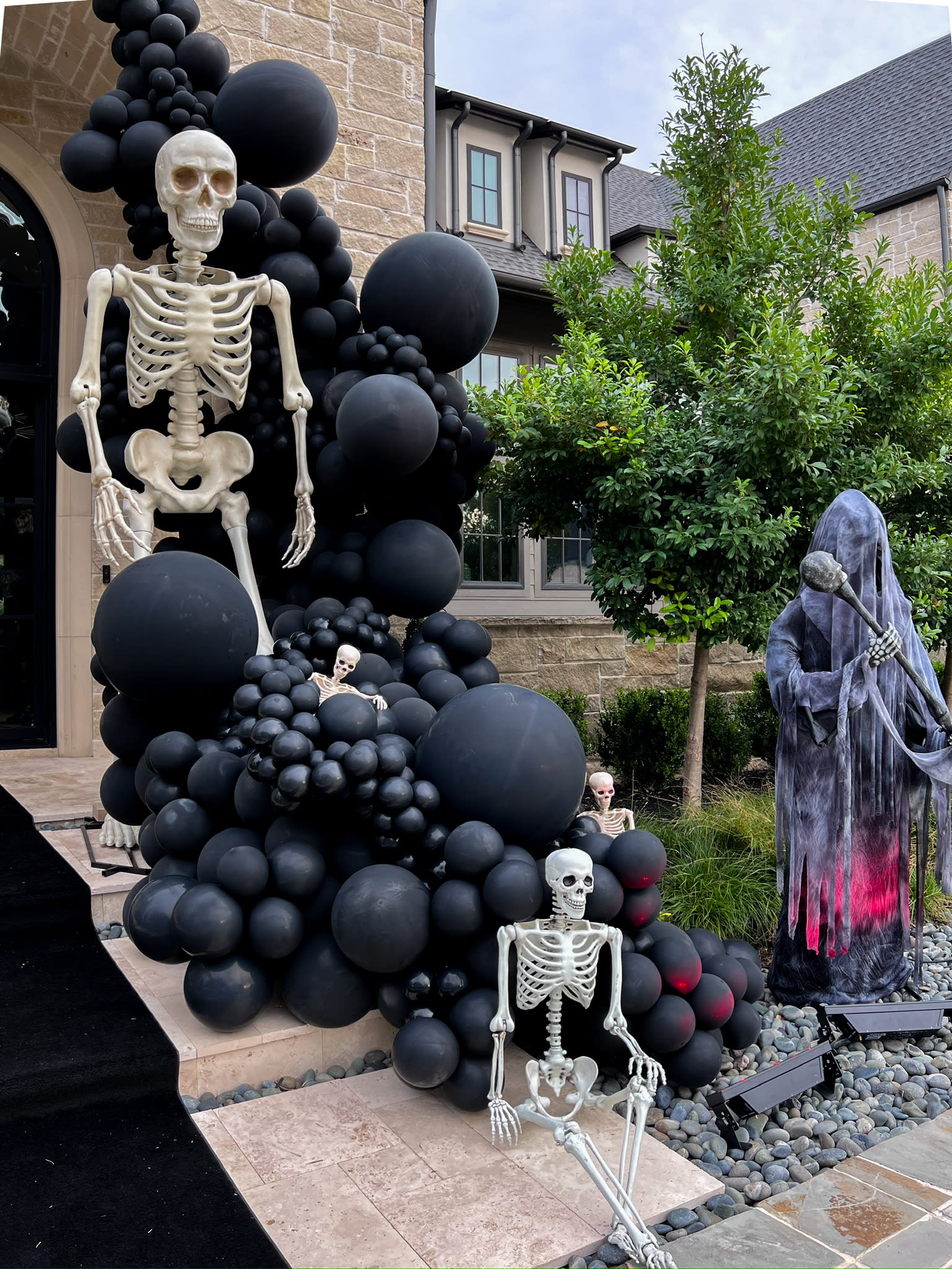 A spooky Halloween display featuring a large skeleton figure surrounded by black balloons and a hooded figure, set against the backdrop of a brick building with greenery.