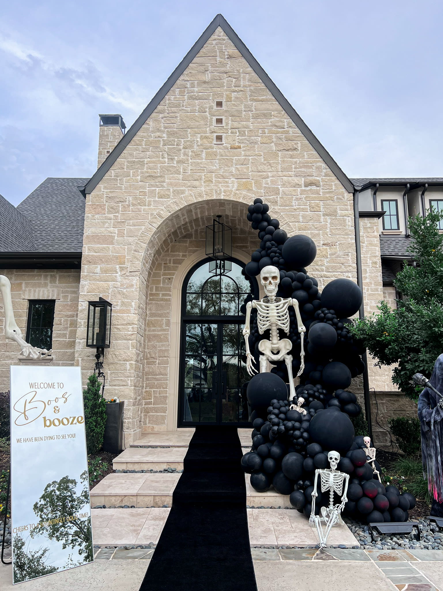 A large, stone-built structure with a prominent arched entrance, surrounded by an elaborate Halloween-themed display featuring a large skeleton figure and numerous black balloons.